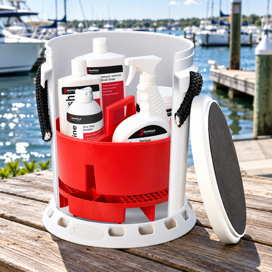 A white and red outdoor cleaning organizer bucket with black handles sits on a wooden dock with a marina and boats in the background. Inside the bucket are several white bottles and containers, and a round black-and-white filter or pad leans against the bucket on the right.
